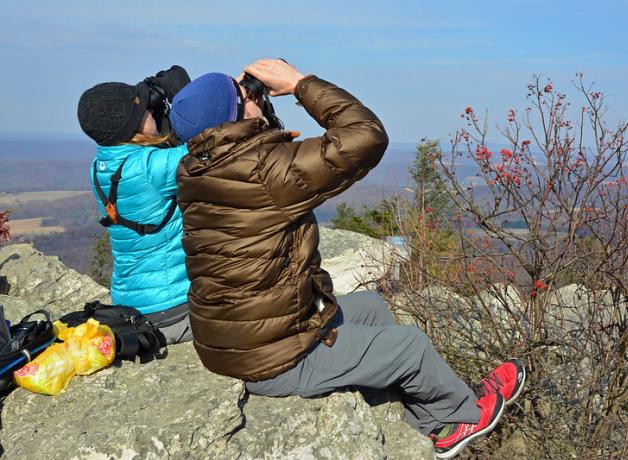 Two people sitting on rocky outcropping and looking up through binoculars on a sunny day. 