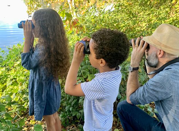 A little girl and boy with their father on a leafy bank, each using binoculars looking out toward the water