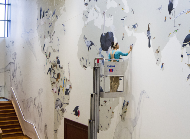 A photo of Jane Kim painting the Wall of Birds mural in a lift several feet off the ground.