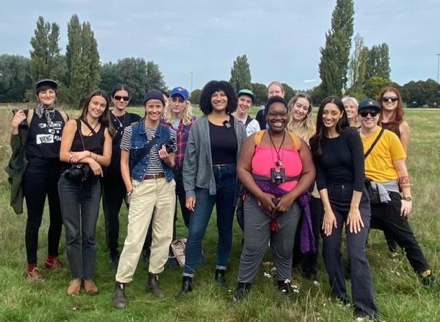 A group of birders, some holding binoculars, pose together in a field, smiling at the person taking the photo