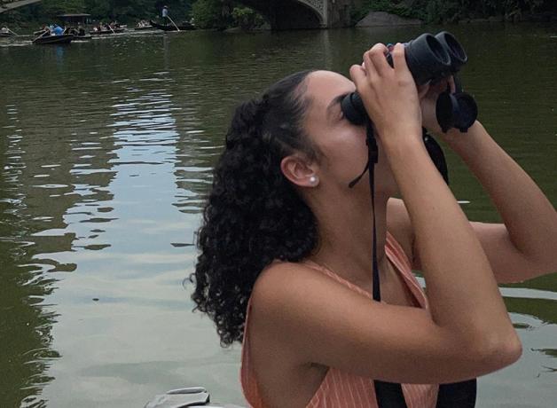 Haley Scott holding binoculars to her eyes as she looks upward while birdwatching from a boat.