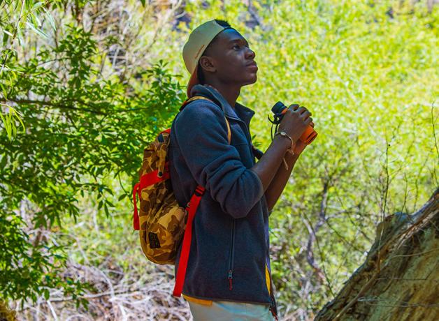 A young Black man is outdoors amidst greenery in filtered sunlight, while holding binoculars and wearing a backpack. 