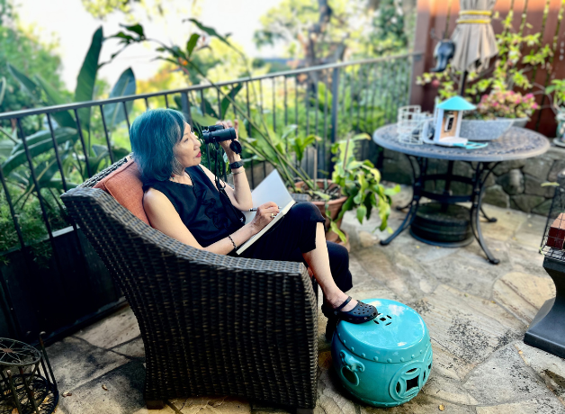 Amy Tan sits in a chair in her backyard, holding binoculars up to her face while writing in a notebook