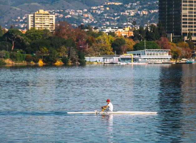 A view of Lake Merrit, with a man rowing across the water in the foreground, and trees and buildings reflecting on the water from the shore.