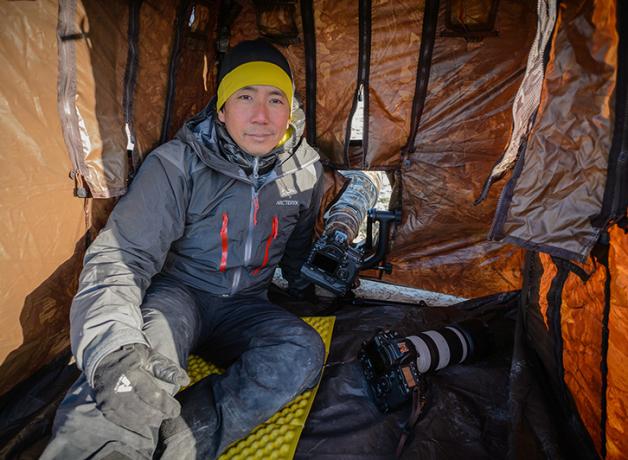 Photographer and author Noppadol Paothong, dressed for cold weather, sits inside a tent with openings for his cameras used for photographing wildlife.