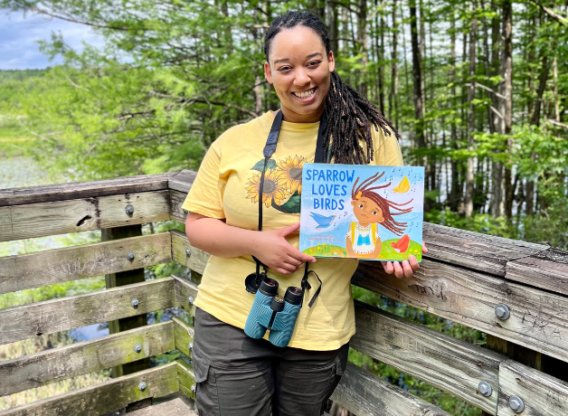 A photo of Murry Burgess standing outside with binoculars and a copy of her children's book.