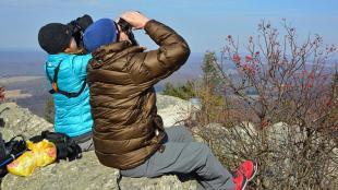 Two people sitting on rocky outcropping and looking up through binoculars on a sunny day. 