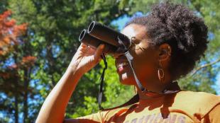 Dara Wilson in a yellow shirt looks through binoculars while standing in sunlight with trees in the background