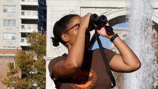 Georgia Silvera Seamans, a Black woman, raises binoculars to her face while sitting in sunlight near a fountain at the Washington Square Park in Manhattan. Georgia is the director of an organization called Washington Square Park Eco Projects.
