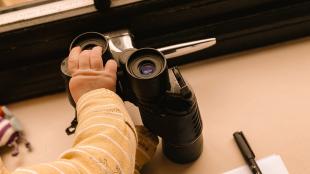 A baby's hand rests on a pair of binoculars that are on a windowsill. A pen and piece of paper are to the right of the binoculars.