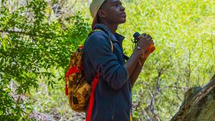 A young Black man is outdoors amidst greenery in filtered sunlight, while holding binoculars and wearing a backpack. 