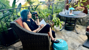 Amy Tan sits in a chair in her backyard, holding binoculars up to her face while writing in a notebook