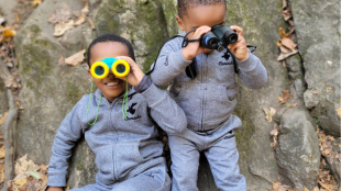 Two little boys in matching outfits holding binoculars outside.
