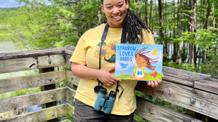 A photo of Murry Burgess standing outside with binoculars and a copy of her children's book.