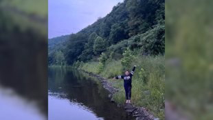 Woman wearing black stands on the edge of a lake, with arms stretched out