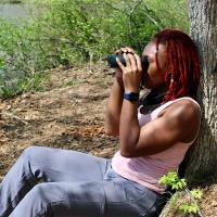 Deja Perkins sitting on the ground in filtered sunlight, with bushes and water in the background. Deja's back is against a tree trunk as she holds binoculars to her eyes, and she's wearing a pink tank top and blue pants.