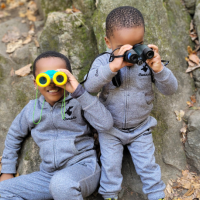 Two little boys in matching outfits holding binoculars outside.
