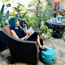 Amy Tan sits in a chair in her backyard, holding binoculars up to her face while writing in a notebook